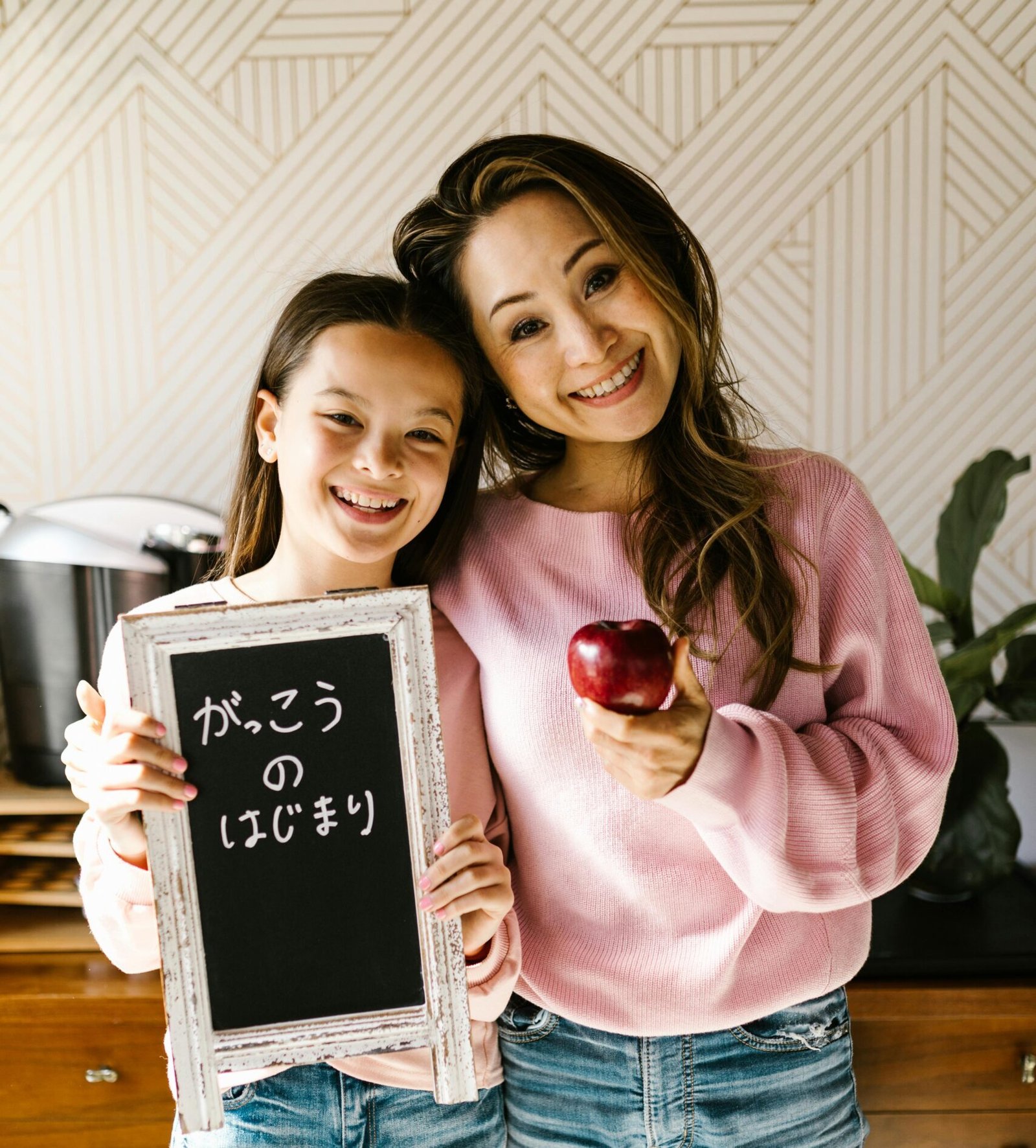 Una madre y su hija de origen japonés sonríen a la cámara en una habitación luminosa. La niña sostiene una pequeña pizarra negra con caligrafía japonesa que anuncia el inicio de clases, mientras la madre sostiene una manzana roja. Ambas visten suéteres rosas y jeans, transmitiendo un ambiente de felicidad y educación en el hogar.